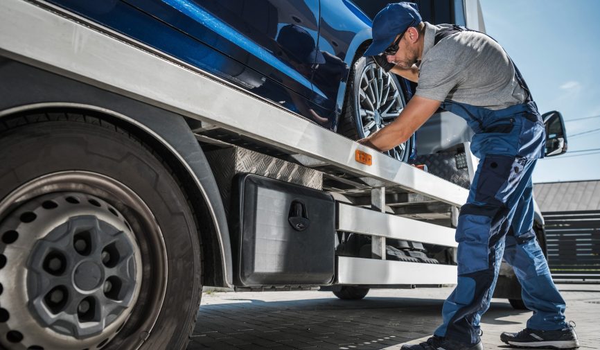 Tow Truck Driver Preparing to Safely Deliver Brand New Car From Dealer to Buyer. Vehicle Transportation Theme.