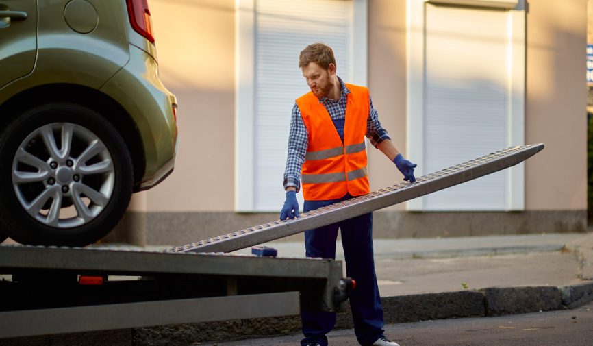 Worker preparing tow truck platform for car Male road worker in uniform preparing tow truck platform for car loading on city street side
